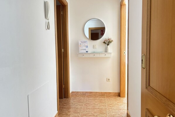 Un pasillo con una puerta de madera, un espejo, una pequeña estantería con un jarrón y un cartel.