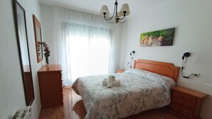 Un dormitorio con cama, mesita de noche, cómoda, radiador, ventana con cortinas, un cuadro y una lámpara de araña.