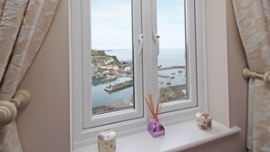 A window with a view of a coastal town, a shelf with decorative items, and curtains. - Wheal Kendall (St Austell)