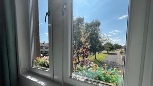 Ein Fenster mit Blick auf eine Wohnstraße, einen Garten mit bunten Blumen und einen klaren Himmel.