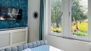 A bedroom with a TV mounted on the wall, a window with a view of a garden, and a bed with a patterned bedspread. - Entire Pet Friendly Apartment- Self Check In/Out (Kelty)