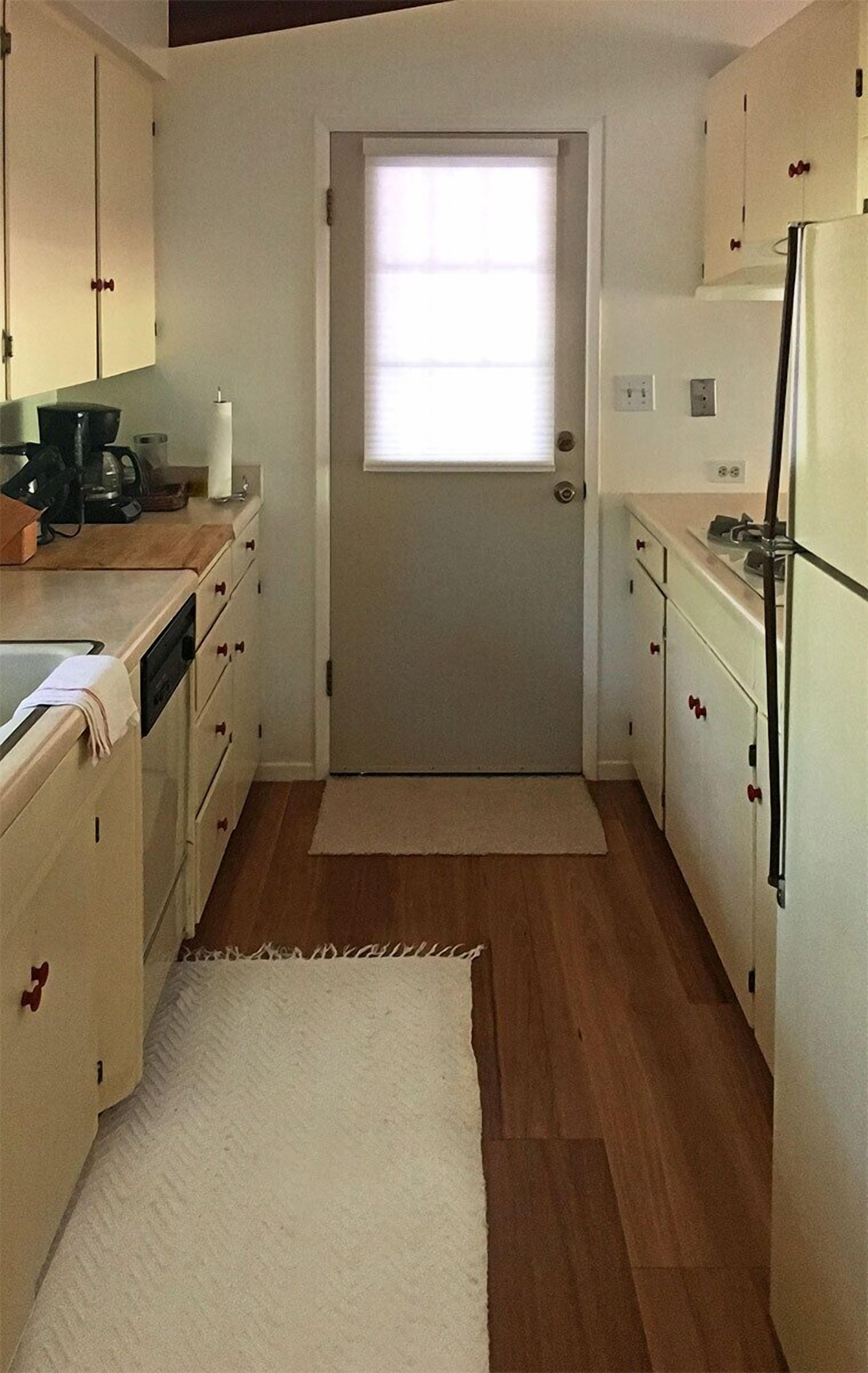 A compact kitchen with wooden flooring, white cabinets, a refrigerator, and a window with blinds.