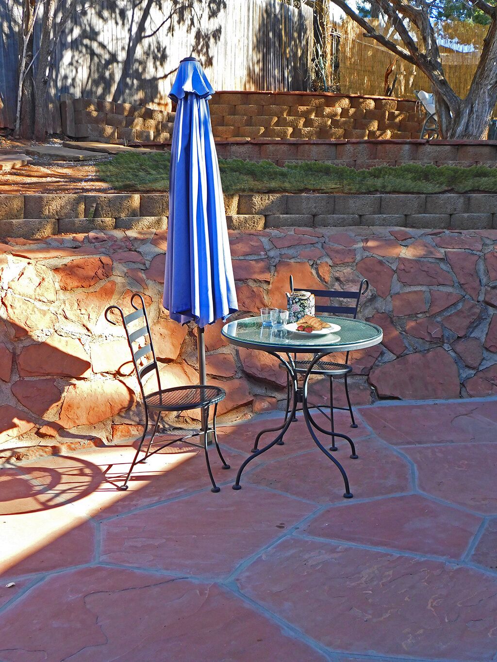 Eine Terrasse mit rundem Tisch und zwei Stühlen, einem blauen Sonnenschirm und einer Steinmauer im Hintergrund.