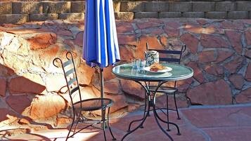 Eine Terrasse mit rundem Tisch und zwei Stühlen, einem blauen Sonnenschirm und einer Steinmauer im Hintergrund.
