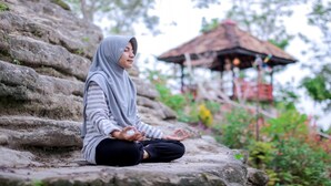 A person sitting cross-legged on a stone ledge outdoors, with a thatched-roof structure in the background. - Camping Ground Gunung Mijil Hils (Bantul)