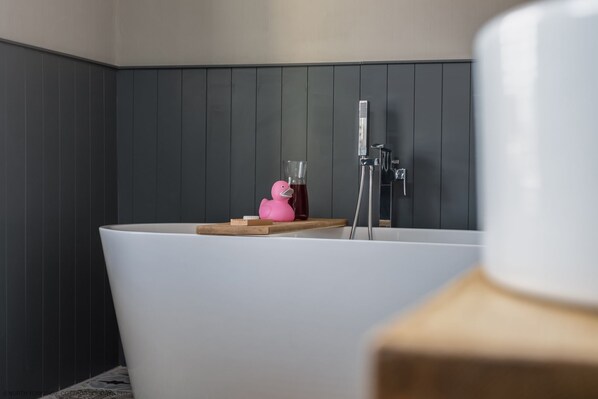A modern bathroom with a freestanding bathtub, a wooden shelf with a pink rubber duck and a glass of red liquid, and a shower with a handheld showerhead.