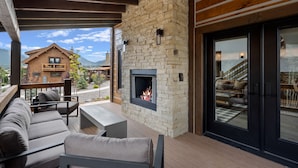 A covered outdoor seating area with a stone fireplace, wooden deck, and a view of a mountain and a lodge. - Elk Haven! Private Jacuzzi, Indoor/Outdoor Fireplace, Two Large Patios (Estes Park)