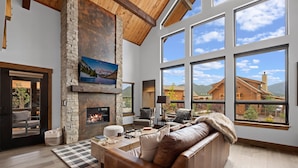 A spacious living room with a stone fireplace, large windows, and a view of a mountain cabin. - Elk Haven! Private Jacuzzi, Indoor/Outdoor Fireplace, Two Large Patios (Estes Park)