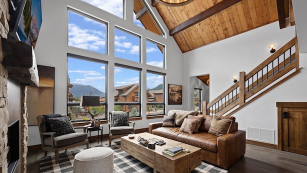 A spacious living room with a large window, a fireplace, a leather sofa, and a wooden staircase. - Elk Haven! Private Jacuzzi, Indoor/Outdoor Fireplace, Two Large Patios (Estes Park)