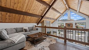 A spacious living room with a high wooden ceiling, a gray sofa, a wooden coffee table, and a large window overlooking a scenic landscape. - Elk Haven! Private Jacuzzi, Indoor/Outdoor Fireplace, Two Large Patios (Estes Park)
