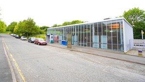 A modern building with large glass windows, a parking lot with several cars, and a green, tree-lined street. - Newly Refurbished Studio, 20mins to London Bridge (Orpington)