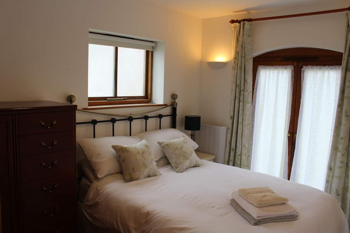 Ein Schlafzimmer mit Bett, Schminktisch, Nachttisch, Lampe und einem Fenster mit Vorhängen.