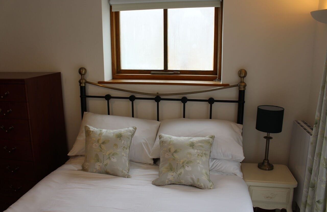 A bedroom with a bed, two floral pillows, a bedside table with a lamp, a wooden dresser, and a window with a wooden frame.