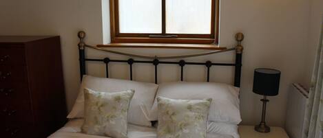 A bedroom with a bed, two floral pillows, a bedside table with a lamp, a wooden dresser, and a window with a wooden frame.