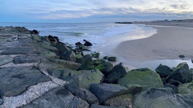 Eine felsige Küstenlinie mit einem ruhigen Strand und Gebäuden in der Ferne.