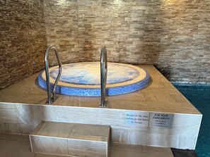A hot tub with a stone wall backdrop, metal steps, and safety signs.