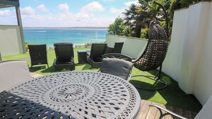 A patio with a round metal table, four chairs, and a hanging chair overlooking a beach. - Peninsula Apartment 2 (St Ives)