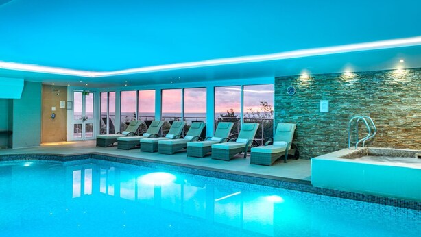 An indoor swimming pool with lounge chairs, a stone wall, and a view of the sunset. - Peninsula Apartment 2 (St Ives)