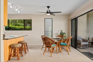 A modern kitchen with a dining area, featuring a ceiling fan, bar stools, and a view of greenery outside.