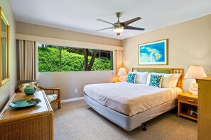 A hotel room with a large bed, a ceiling fan, a window with a view of greenery, and a framed map on the wall.