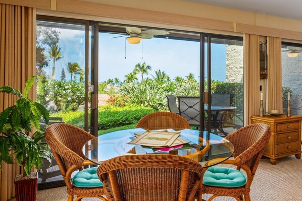 A room with a glass table surrounded by wicker chairs, a wooden cabinet, and a view of a garden with palm trees.