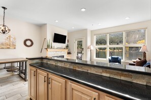A modern kitchen with a stone countertop, wooden cabinets, a flat-screen TV, and a view of a forest through large windows. - Patio access to Keystone Lake & Gorgeous Views, Easy Access to Everything! (Keystone)