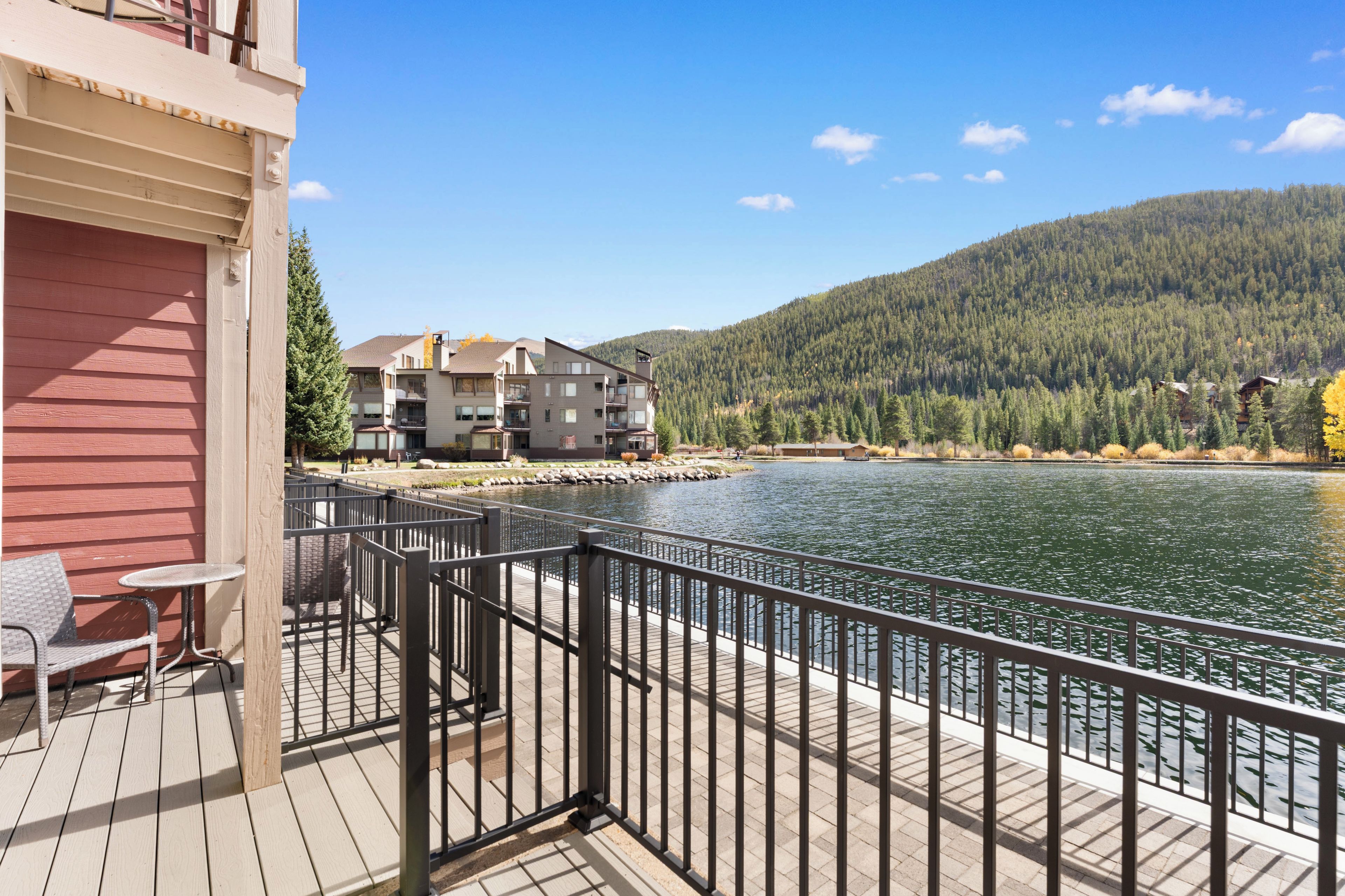 A wooden deck with a metal railing overlooking a calm lake, surrounded by forested hills.