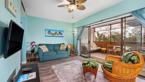 Un salon avec un canapé de couleur turquoise, un fauteuil en osier, un téléviseur à écran plat et un ventilateur de plafond.