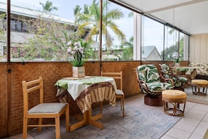 Enjoy a tranquil retreat with a view of palm trees and a building from the large window. The room features a dining table, wicker sofa, and comfortable seating for two.