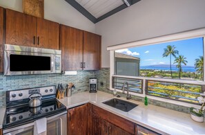 A modern kitchen with wooden cabinets, stainless steel appliances, and a large window overlooking a scenic view. - Wailea Ekolu #604 Ocean View! Walking Distance to Ulua Beach, Wifi, Full A/C (Wailea)