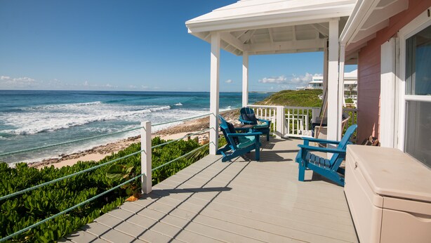 A covered deck with two blue chairs and a beige cabinet, overlooking a beach with waves and a clear blue sky. - Endless Summer - Perfect Getaway near Tahiti Beach (Hope Town)