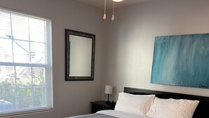 A neatly made bed with a grey comforter and white pillows, a bedside table with a lamp, a framed mirror, and a blue abstract painting on the wall. - Ground-Floor Beach Retreat – Walk to the Sand from This 3BR/2BA Condo (New Smyrna Beach)