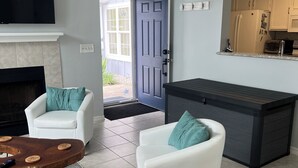 Un salon moderne doté d’une cheminée, de deux canapés blancs agrémentés de coussins turquoise, d’une table basse et d’un coin cuisine avec des placards et des appareils électroménagers.