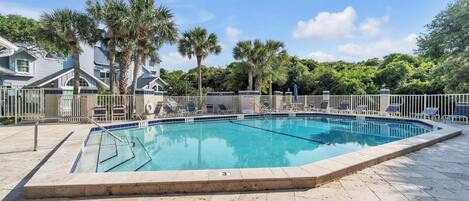 A rectangular swimming pool with a tiled deck, surrounded by palm trees and a white fence.
