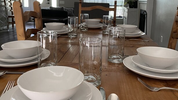 A dining table set with white plates, bowls, and clear glasses, with a wooden floor and a staircase in the background. - Sunrose Cottage - Fresh & Clean - King Suite + free Wifi - Pet Friendly (Cibolo)