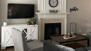 A living room with a fireplace, a mounted TV, a ceiling fan, and a wall-mounted clock. - Sunrose Cottage - Fresh & Clean - King Suite + free Wifi - Pet Friendly (Cibolo)