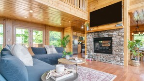 A spacious living area with a blue sofa, a stone fireplace, wooden ceiling, and a kitchen in the background.