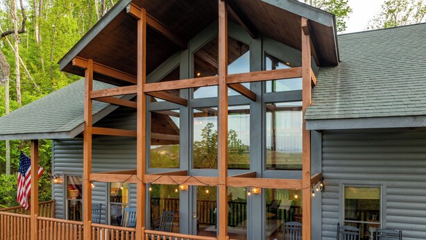 A multi-level wooden cabin with large windows, a deck with outdoor seating, and an American flag.