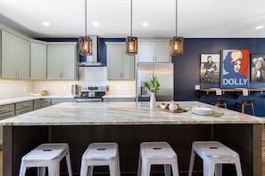 Una cocina moderna con armarios verde claro, una isla de mármol con taburetes, un refrigerador de acero inoxidable e iluminación colgante.