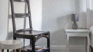 Een hoek van een kamer met een houten stoel, een klein wit tafeltje met een lamp, en een witte muur.