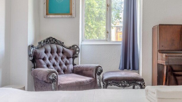 Een kamer met een armstoel met knoopjes, een bijpassend poufje, een houten piano en een raam met uitzicht op groen.