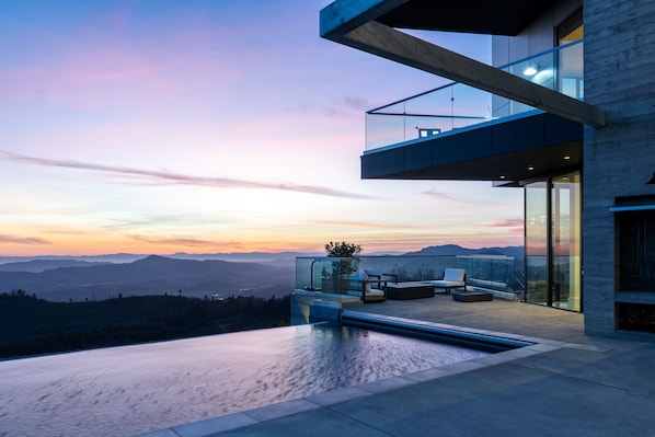 A modern outdoor terrace with a pool, seating area, and a glass balustrade overlooking a mountainous landscape.