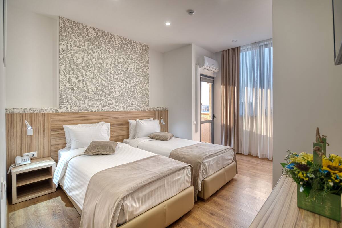 Habitación de hotel con dos camas, cabecera de madera, mesita de noche con teléfono y ventana con cortinas.