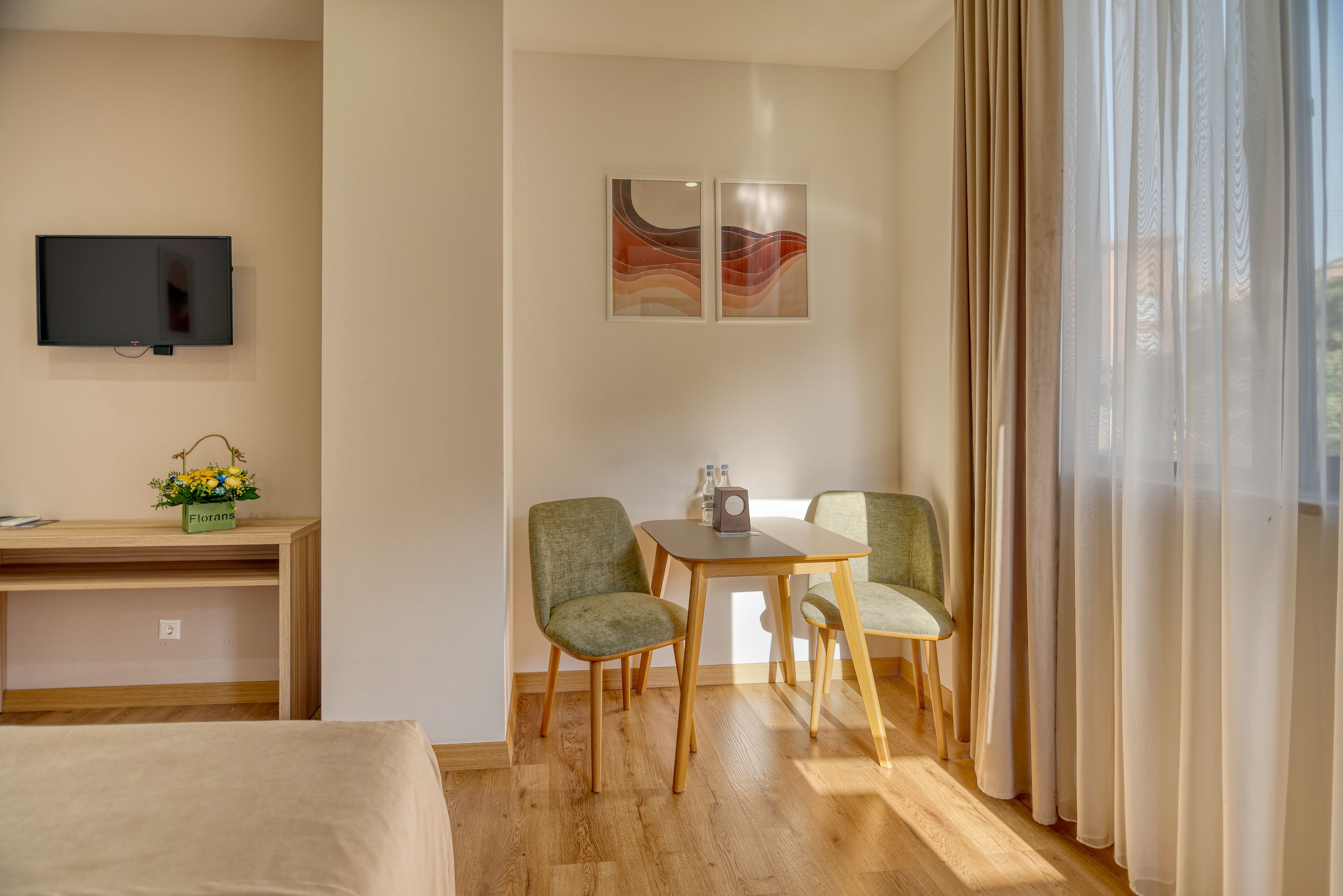Ein Hotelzimmer mit einem Bett, einem Schreibtisch mit Computer, zwei Stühlen um einen Tisch, einem Fernseher an der Wand und einem Fenster mit Vorhängen.