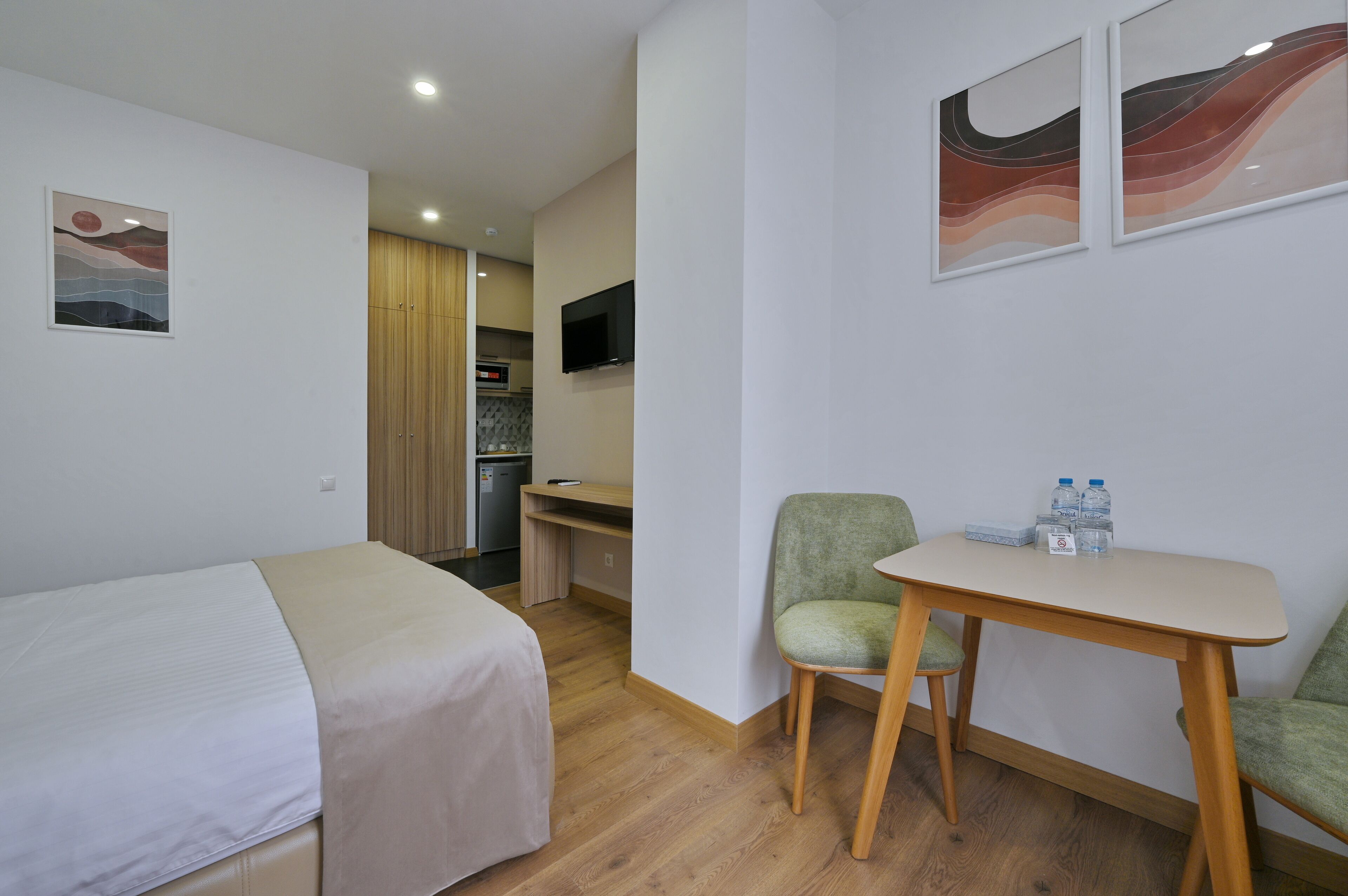 A hotel room with a bed, a desk, a chair, and a small table with water bottles.