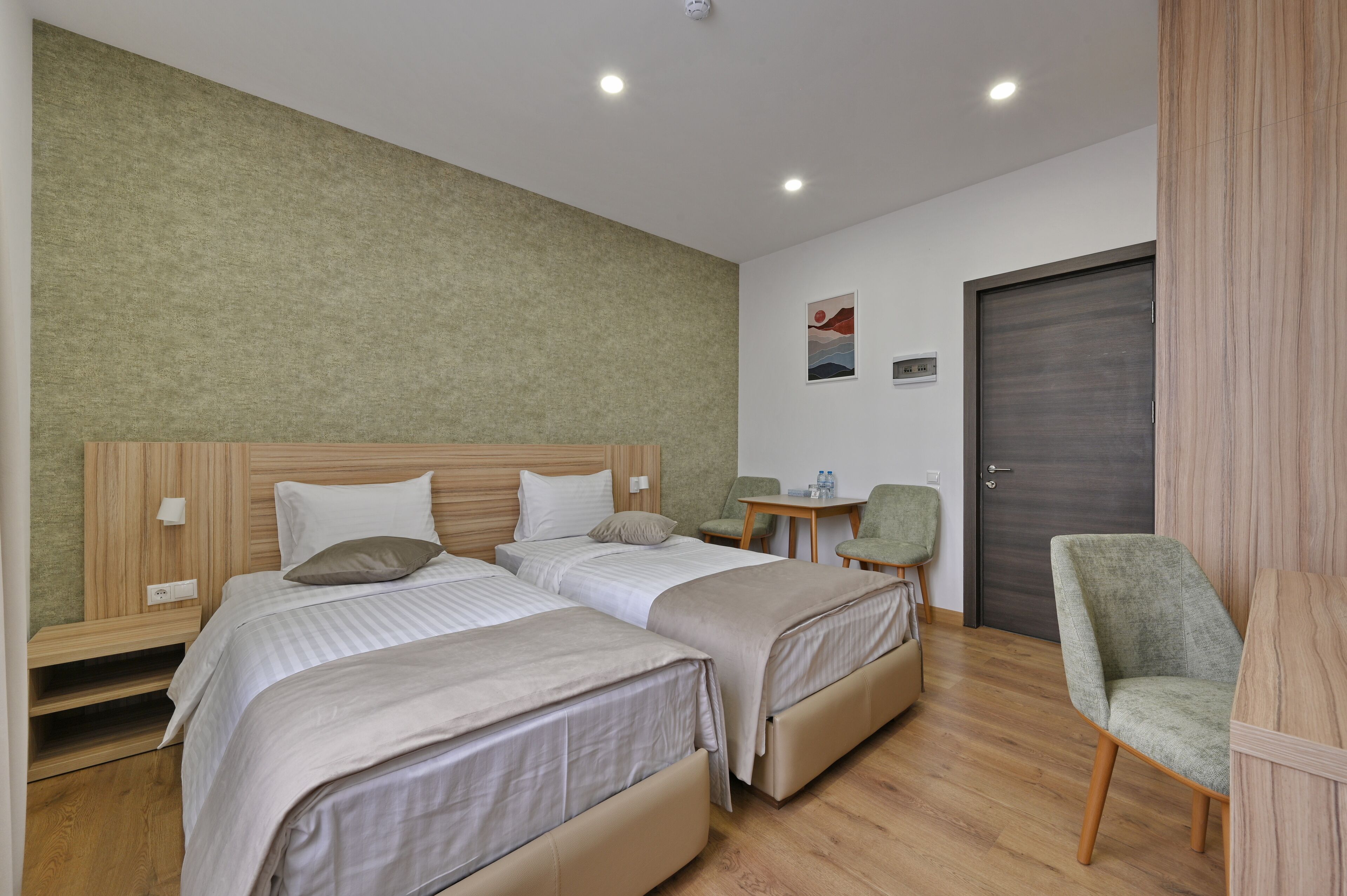 A hotel room with two beds, a small table, a chair, and a door.