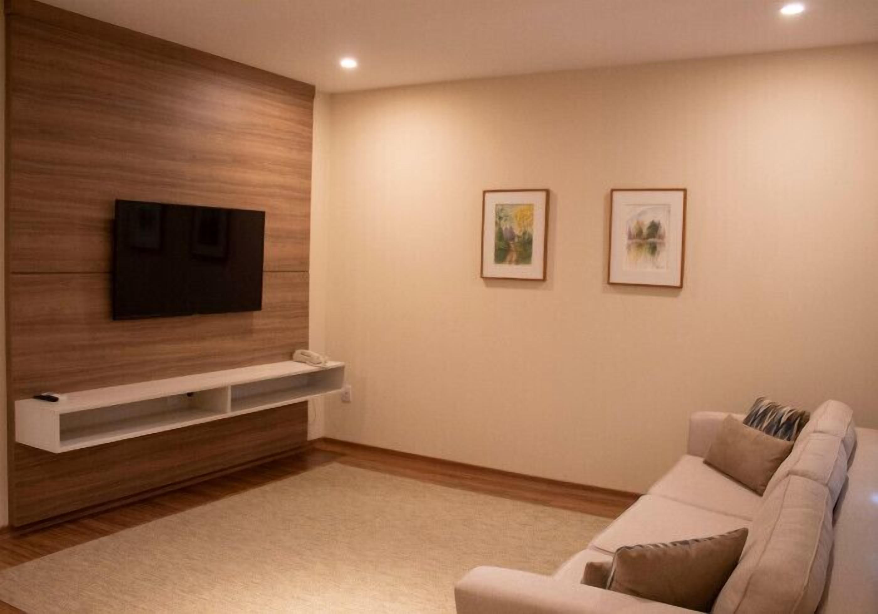 A modern living room with a flat-screen TV mounted on a wooden wall, a white entertainment unit below, a beige sofa with cushions, and two framed artworks on the wall.