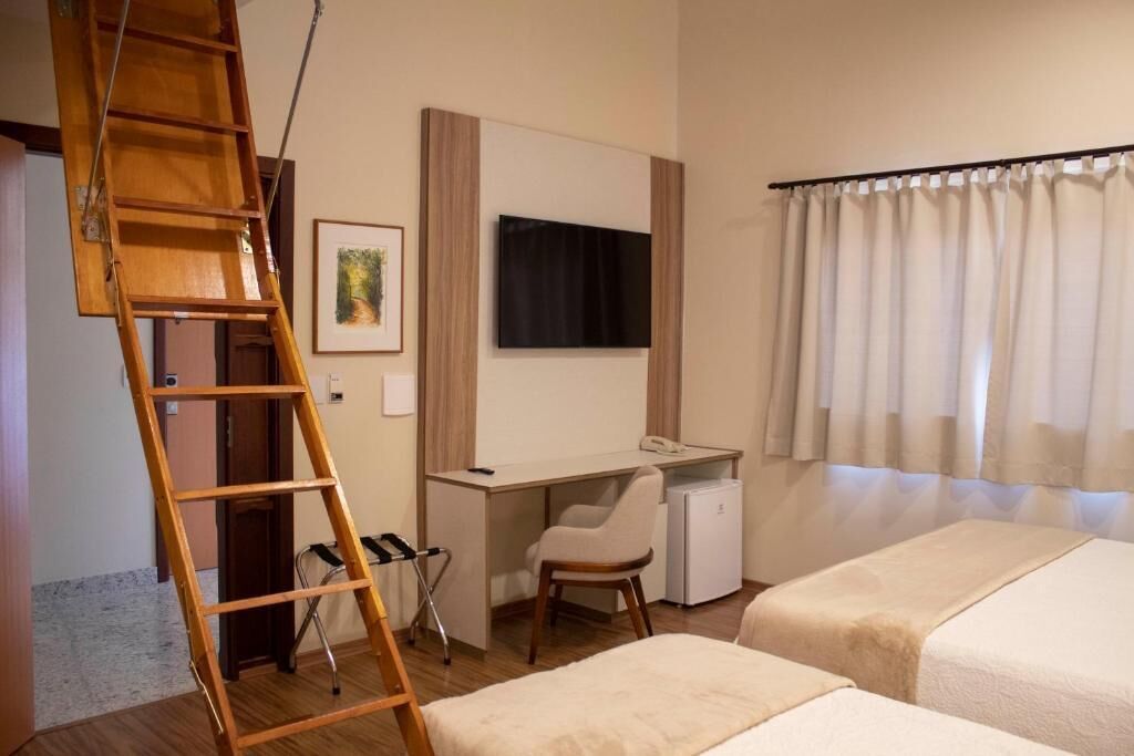 A hotel room with a wooden ladder, a desk with a chair, a bed, a TV mounted on the wall, and a window with curtains.
