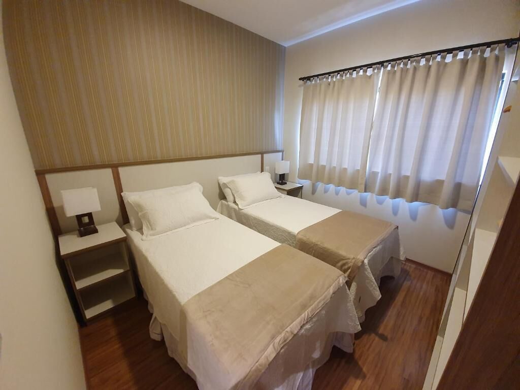 A hotel room with two single beds, a nightstand with a lamp, a wooden headboard, and a window with curtains.
