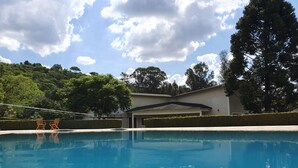 Una piscina con dos sillas de madera, un porche cubierto y un edificio con un garaje al fondo.
